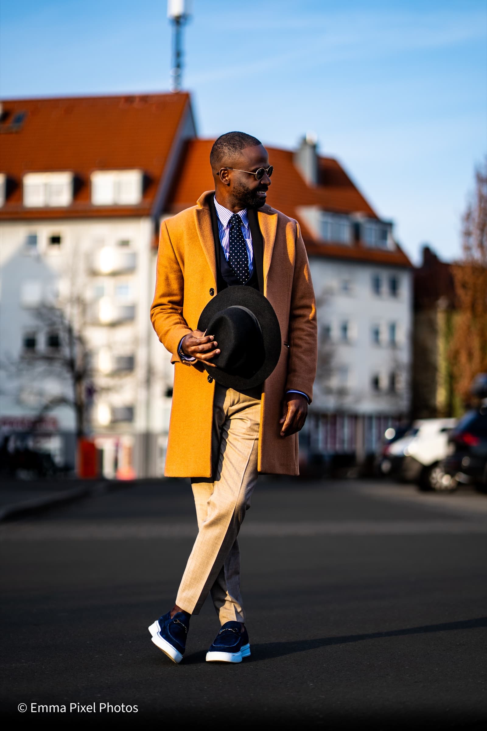 Full-body walking shot in mustard coat, holding fedora