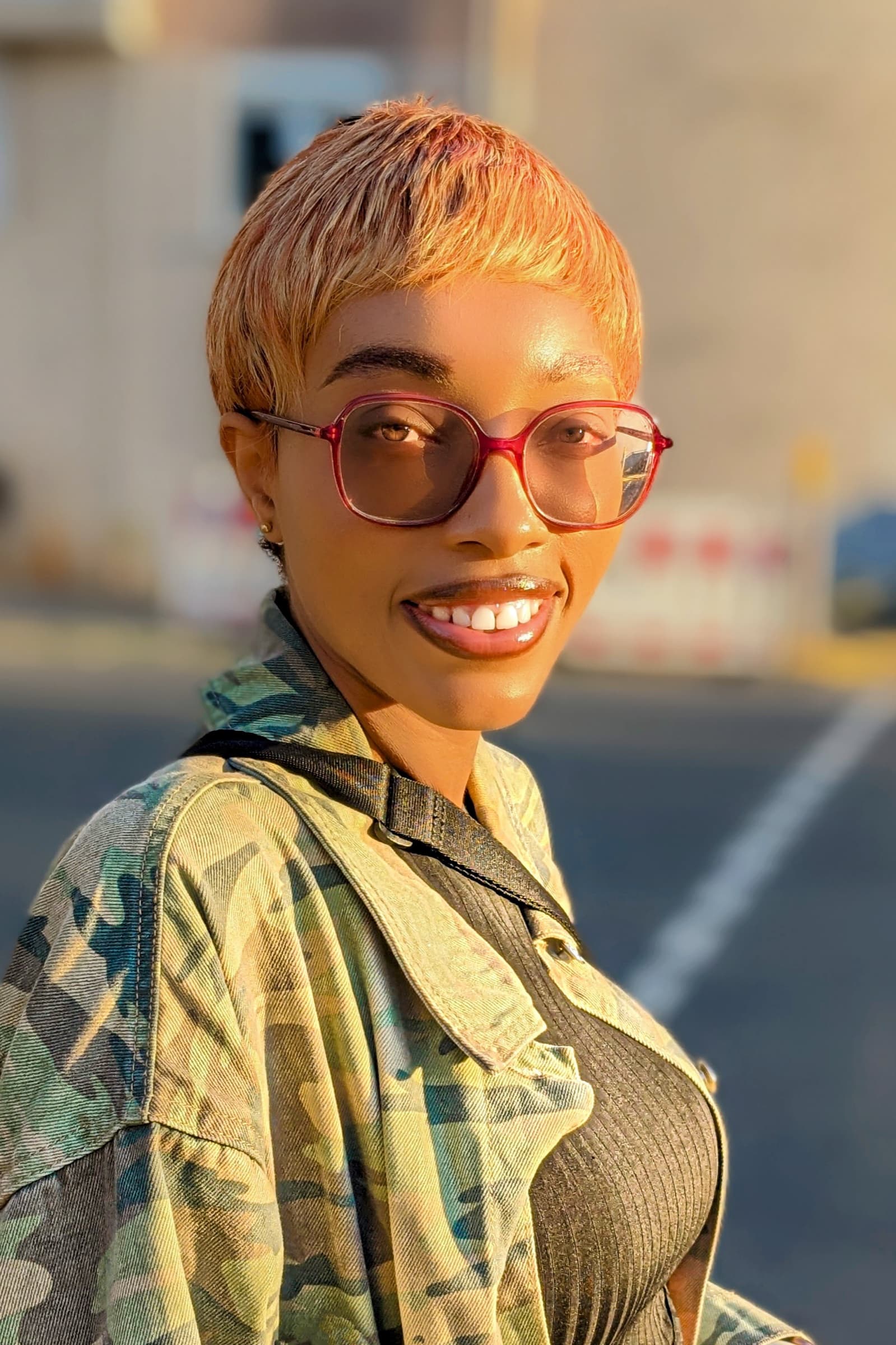 Pixie cut, red sunglasses, camo jacket — golden hour smile